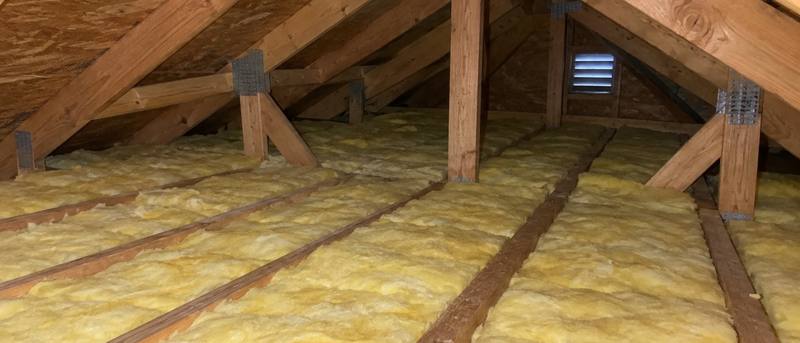 Yellow fiberglass batts laid between attic joists, with a metal gable vent and sunlight on the roof boards