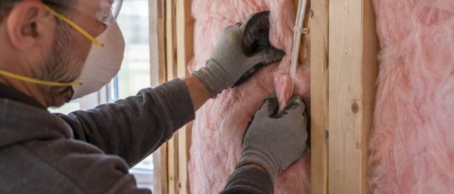 Worker in a dust mask and gloves pressing a pink batt into a wall stud bay beside a window frame