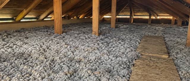 Grey blown cellulose piled deep on both sides of a plywood walkway between roof trusses