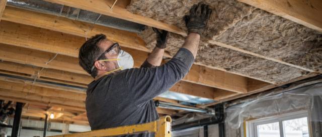 Worker standing on a scaffold pressing a mineral wool batt between ceiling joists