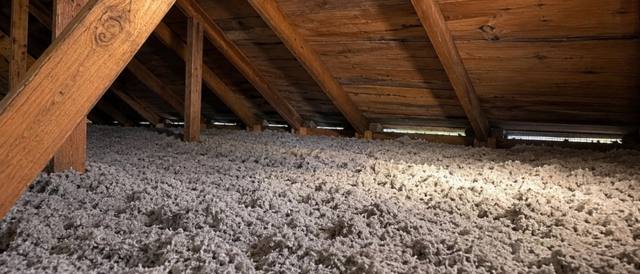 Thick grey-brown blown cellulose covering an attic floor beneath dark roof boards